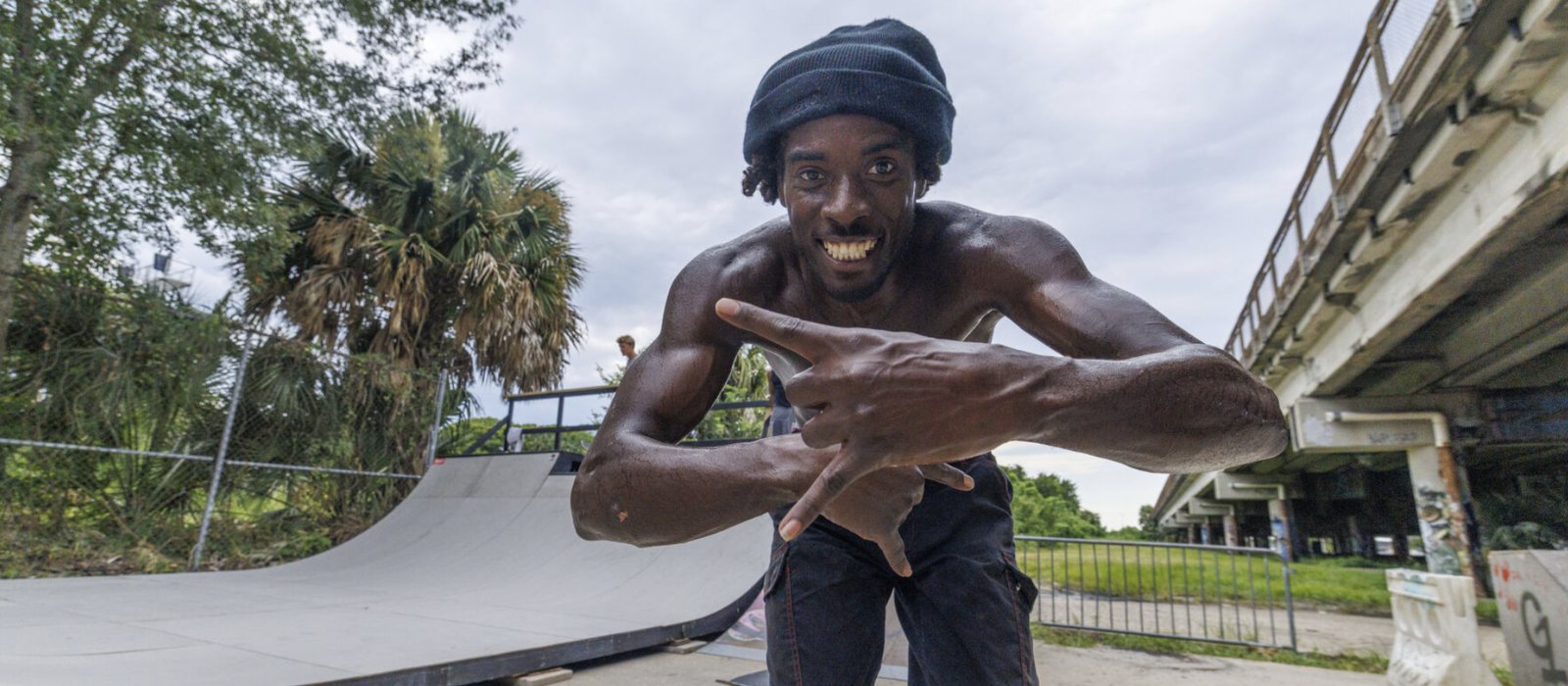 A skater grins at the camera in front of a ramp at a DIY skatepark under an overpass