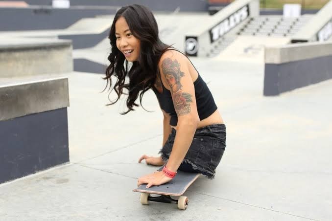 An adaptive skater smiles as she rides a skateboard across a skatepark